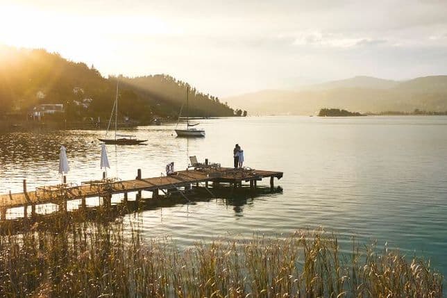 Calm lake in warm backlight; long wooden jetty with white parasols leads into the water, with two people standing at the end. Sailboats, hills and reeds in the foreground.