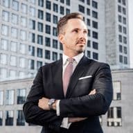 Benjamin Rogmans in a suit stands confidently with arms crossed, in front of modern office buildings under a clear sky.