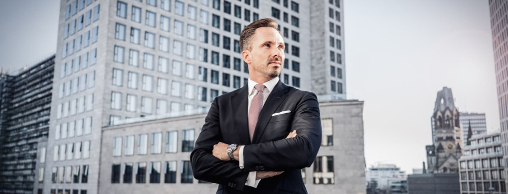 Benjamin Rogmans in a suit stands confidently with arms crossed, in front of modern office buildings under a clear sky.