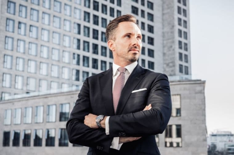Benjamin Rogmans in a suit stands confidently with arms crossed, in front of modern office buildings under a clear sky.