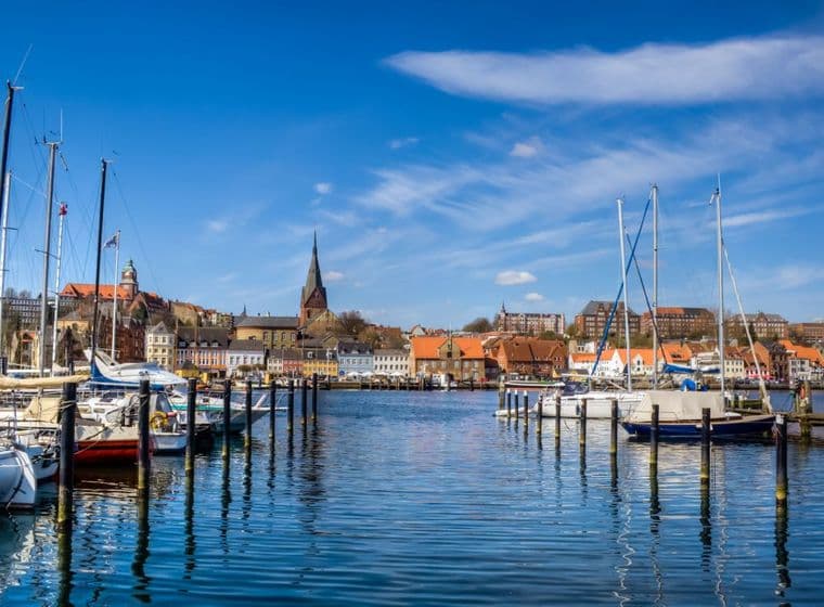 A scenic harbor with sailboats docked, colorful buildings, and a church steeple under a clear blue sky.