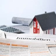Eine kleine Hausmodell-Figur auf einem Zeitungsartikel mit der Überschrift "Immobilien", umgeben von Bauplänen und einem Taschenrechner.