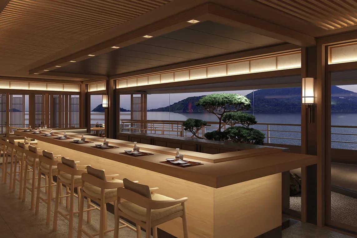 Interior of a warm wooden Japanese-style restaurant with a long counter and stools, bonsai trees by a glass wall overlooking a lake and mountains.