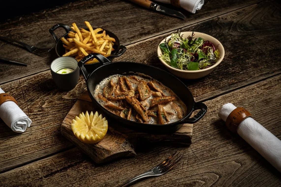 Rustic wooden table set with a cast-iron skillet of fish in sauce, a side of fries, a leafy salad, and a carved pineapple flower on a wooden board.