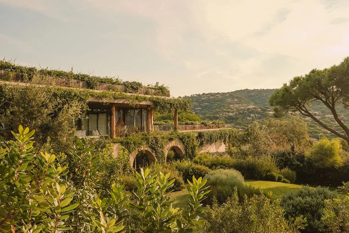 Hillside villa with ivy-draped arches and a glass-walled terrace, surrounded by a lush Mediterranean garden and distant hillside homes.