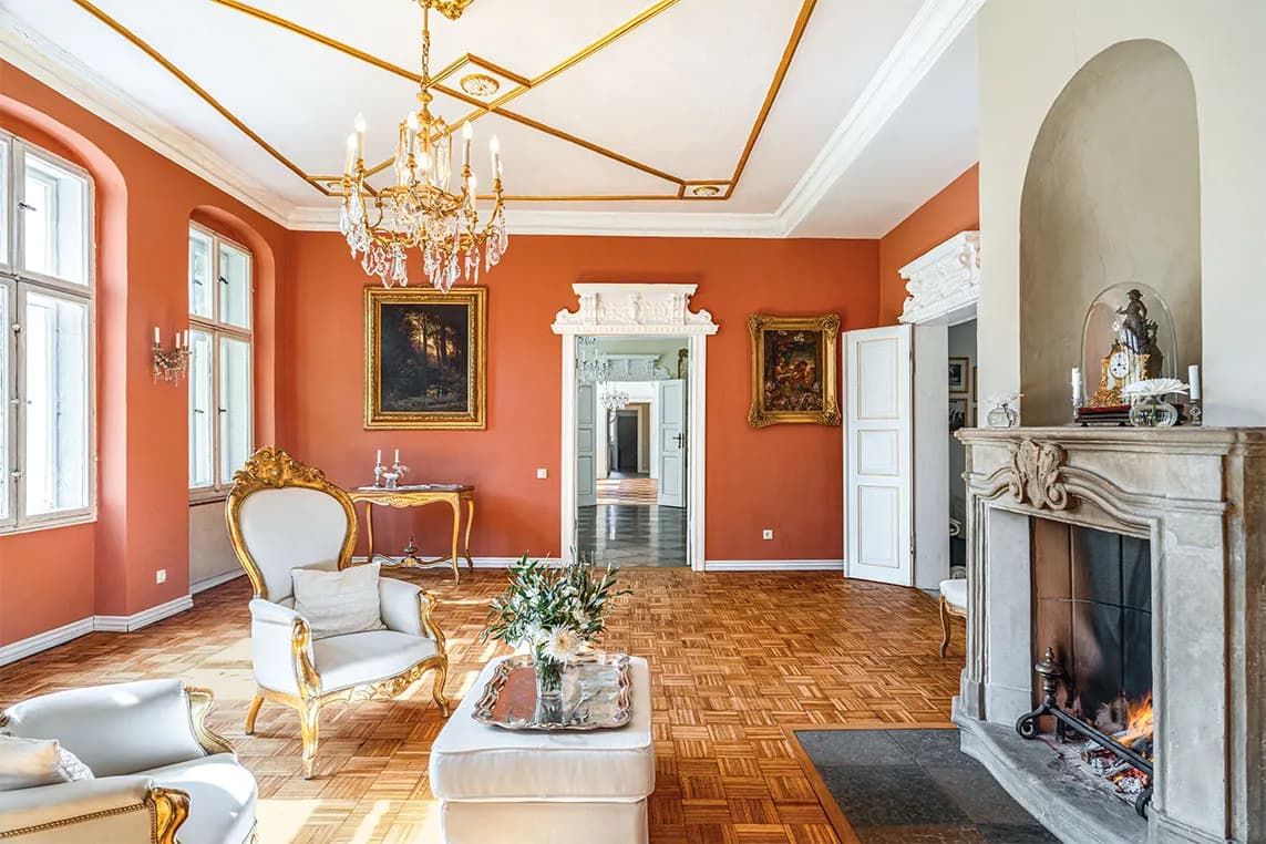 Elegant living room with orange walls, gilded chairs, crystal chandelier, marble fireplace, parquet floor, and framed paintings.