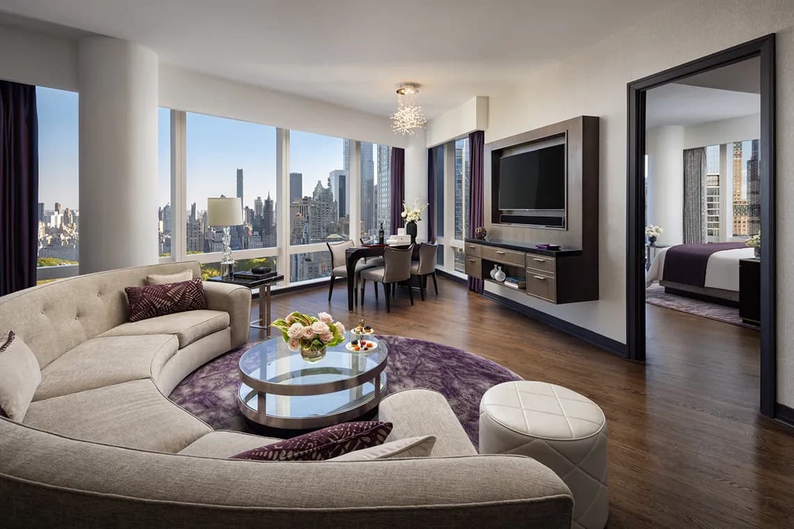 Luxurious living room with curved beige sofa, glass coffee table, purple rug, and city skyline view through large windows; dining area and bedroom doorway.