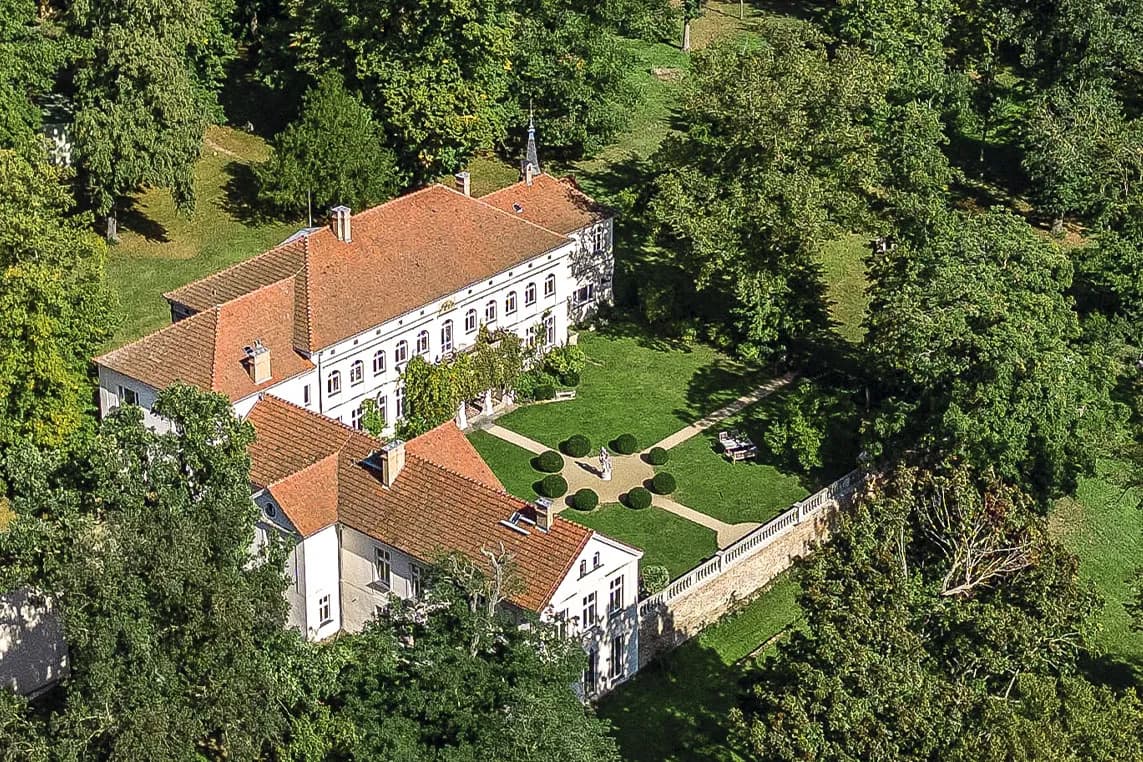 A white mansion with red-tiled roofs sits among lush trees, a manicured lawn with circular shrub beds, and a stone perimeter wall.