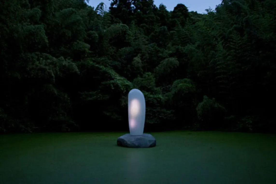 A luminous, oval-shaped sculpture on a rock in a dark green pond, softly glowing amid a dense forest at dusk.