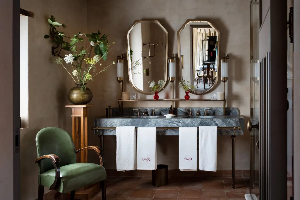 Elegant vintage bathroom with a marble double-sink vanity, twin oval mirrors, brass fixtures, a vase of flowers, and a green upholstered chair; white towels hang below.