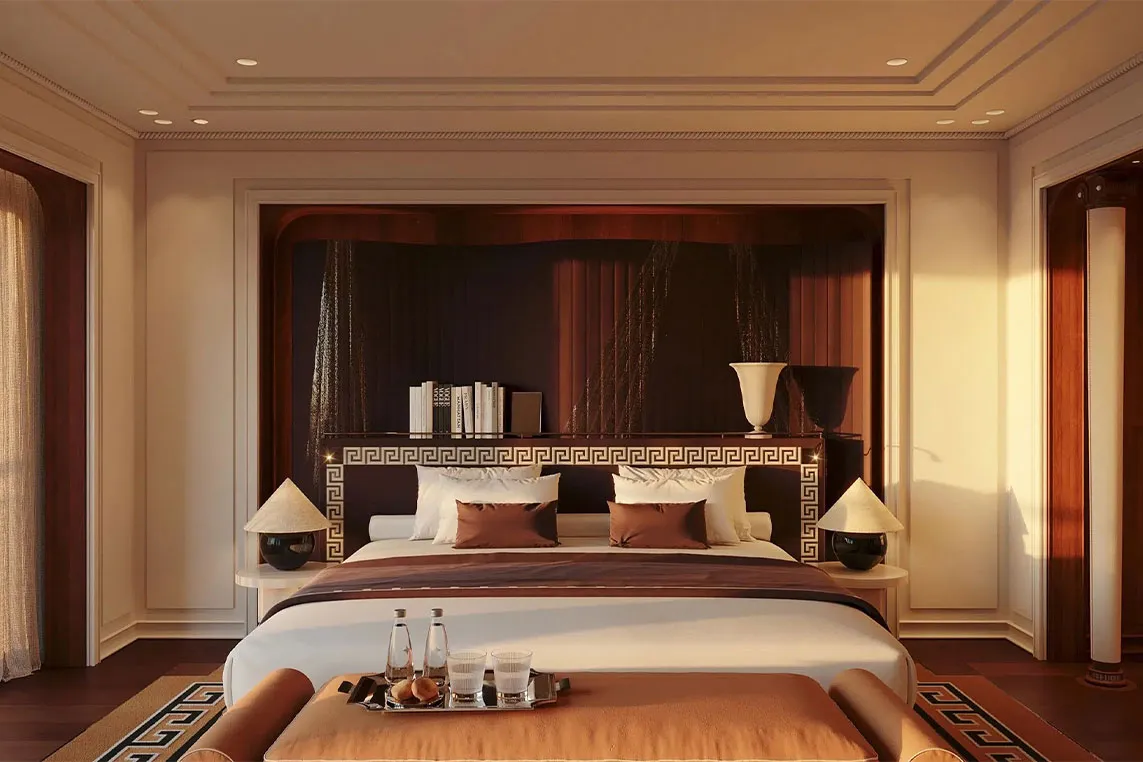 Luxurious hotel bedroom with a large bed and geometric headboard, warm lighting, side lamps, a foot bench with a tray of drinks, and books on a shelf.