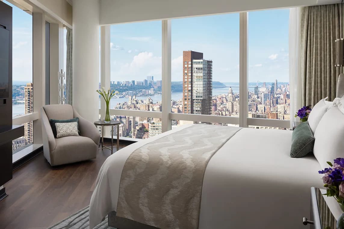 Hotel room with large windows overlooking a city skyline and river; bed with white linens and beige runner, plus a plush chair and flowers.
