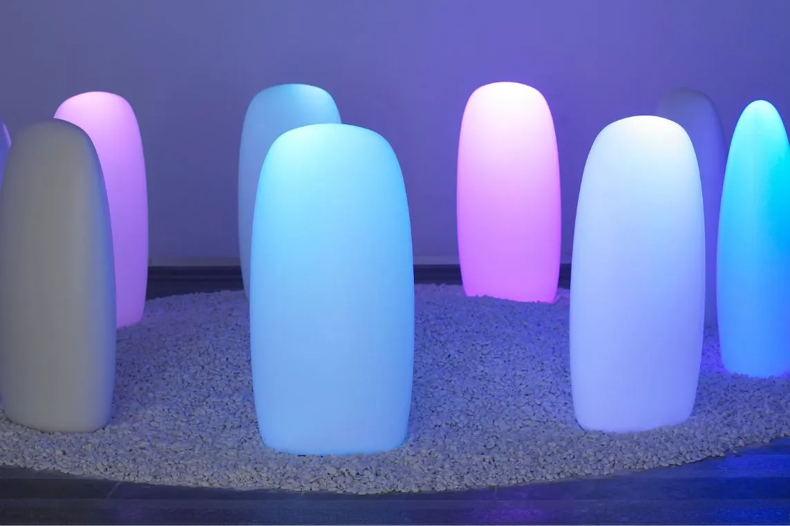 Soft, glowing rounded light sculptures in blue, pink, and purple stand upright on a pebbled circular bed.
