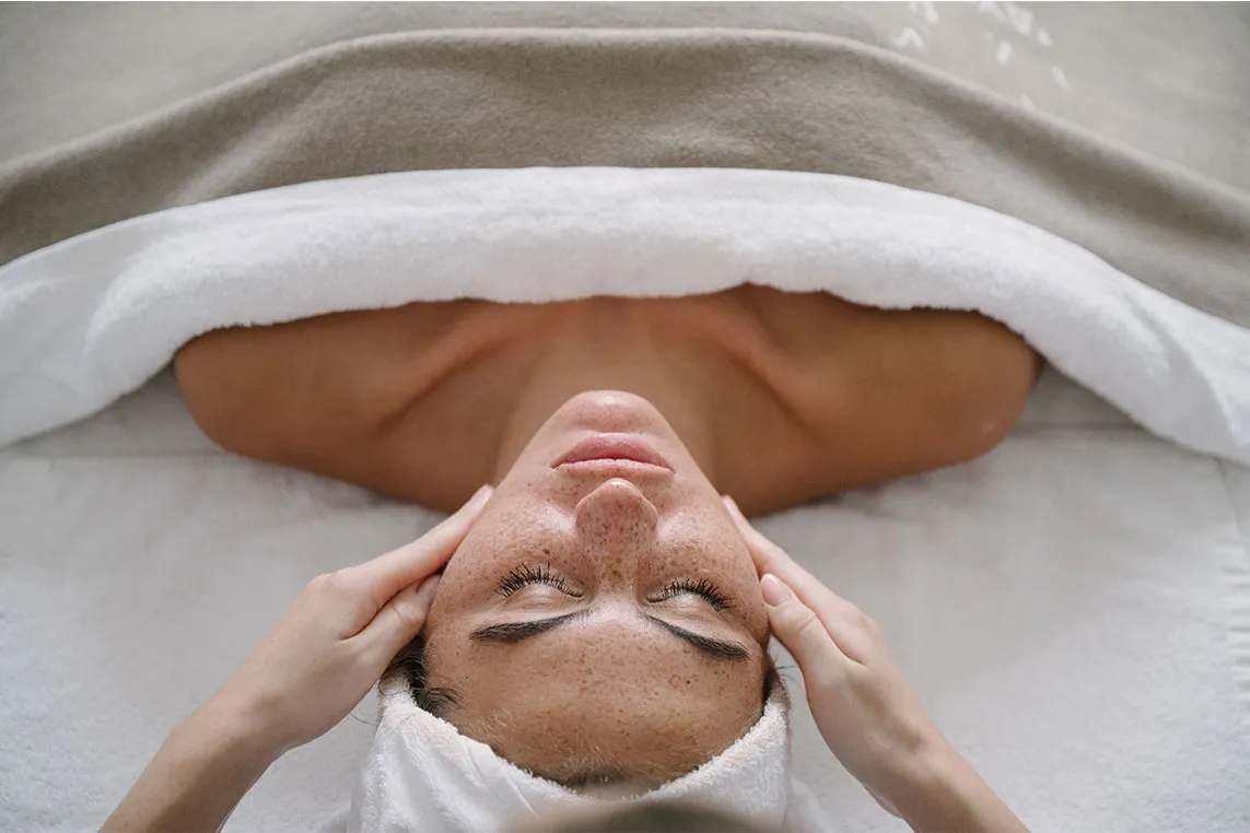 A person lies on a spa bed with a towel wrapped around, eyes closed, as a therapist gently holds their temples for a head massage.