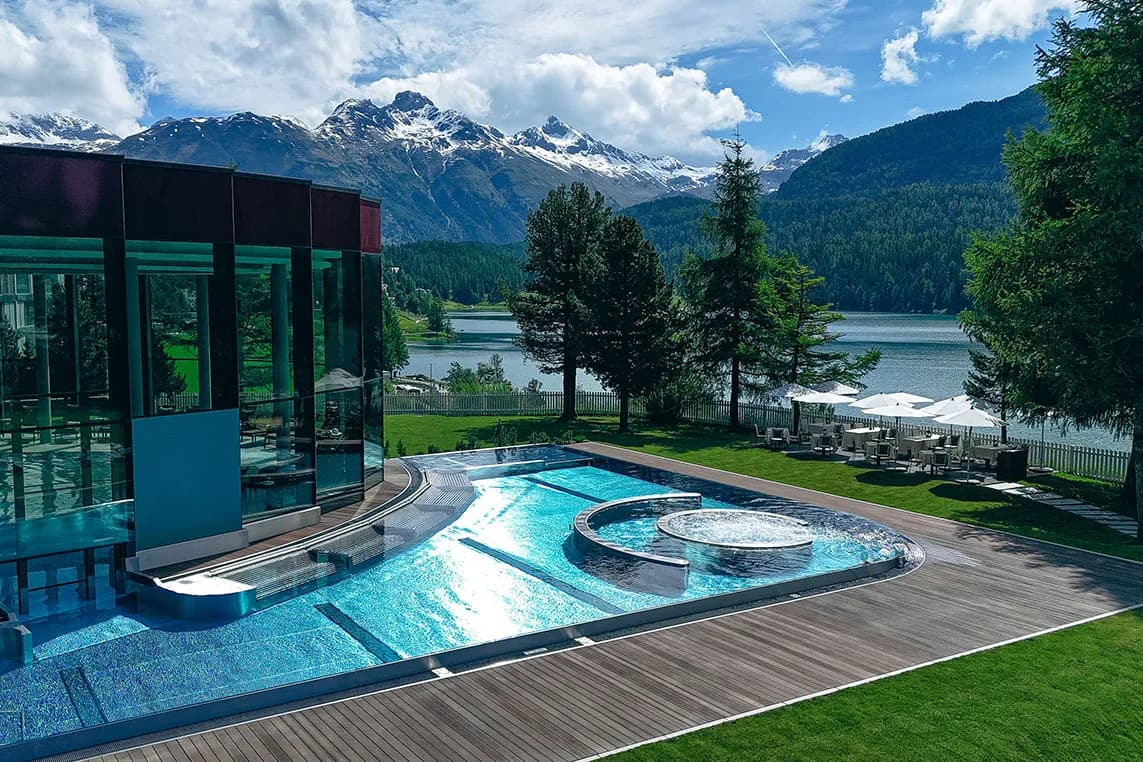 Modern glass building beside a blue pool with a circular spa, wooden deck, lawn, and a lake framed by snow-capped mountains.