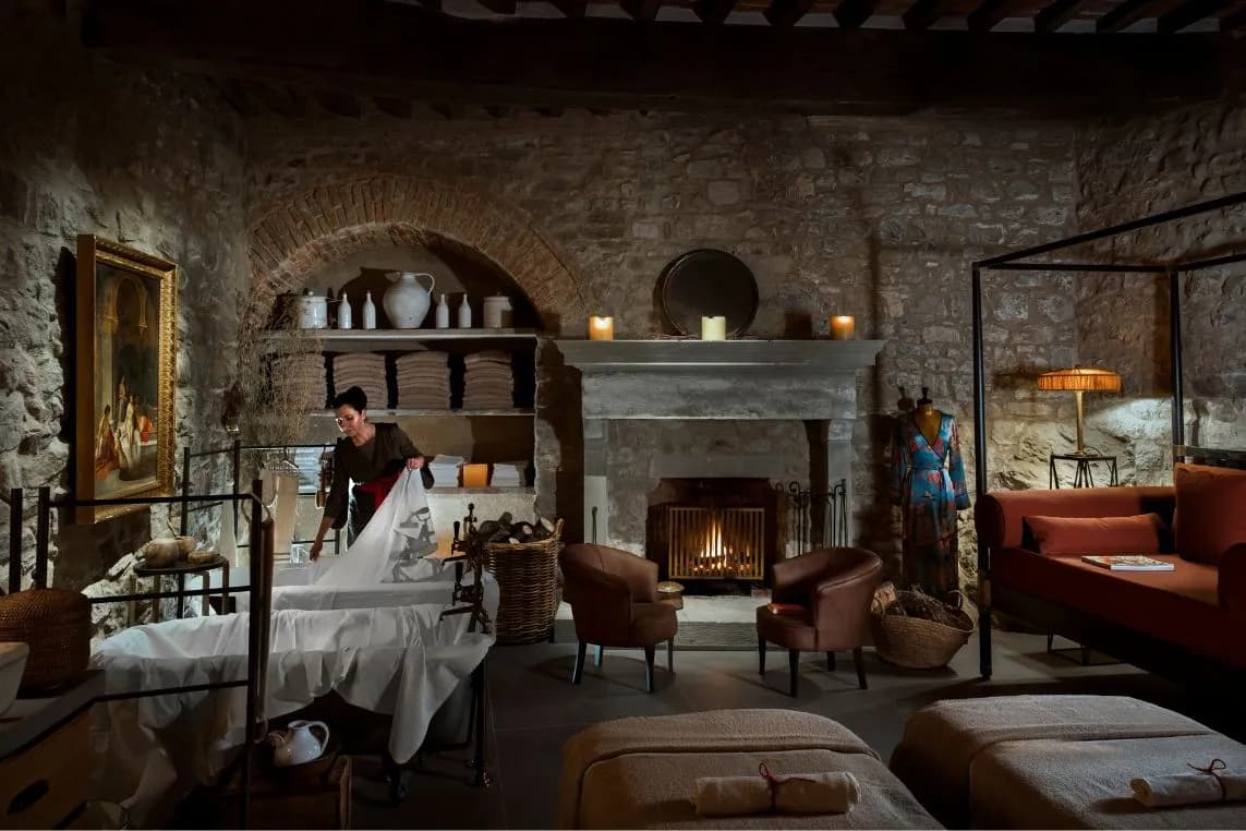 Cozy rustic stone room with a warm fireplace; a person arranges white sheets on a bed, beside a red sofa and shelves with a blue-dress mannequin.