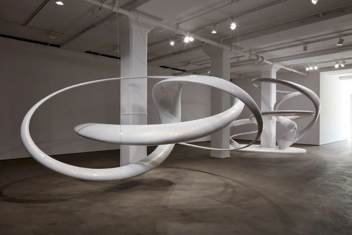 Large white looping sculptures form circular hoops sweeping across a minimalist gallery floor under bright overhead lights.