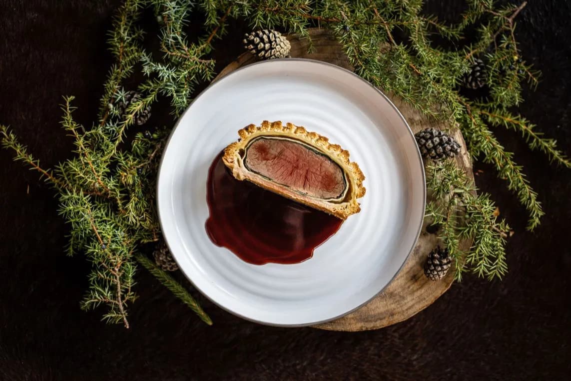 A half-cut pastry pie with red filling on a white plate, with red sauce spilled, placed on a wooden board among evergreen sprigs and pinecones.