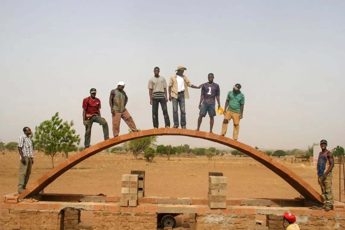 Several men are standing on a large, orange-coloured brick arch that is currently under construction in a barren, arid landscape.
