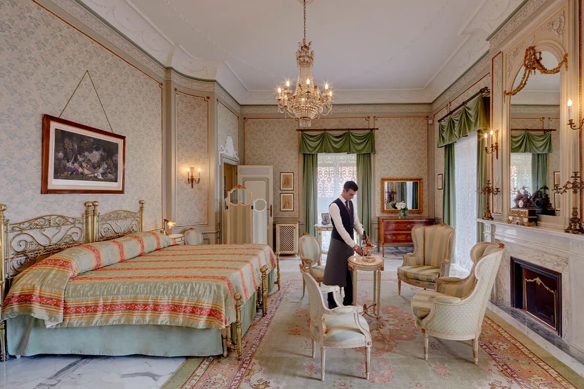 A sumptuous vintage hotel suite featuring a gilded iron bed, a chandelier, green curtains and a waiter serving tea at a small table next to a seating area.