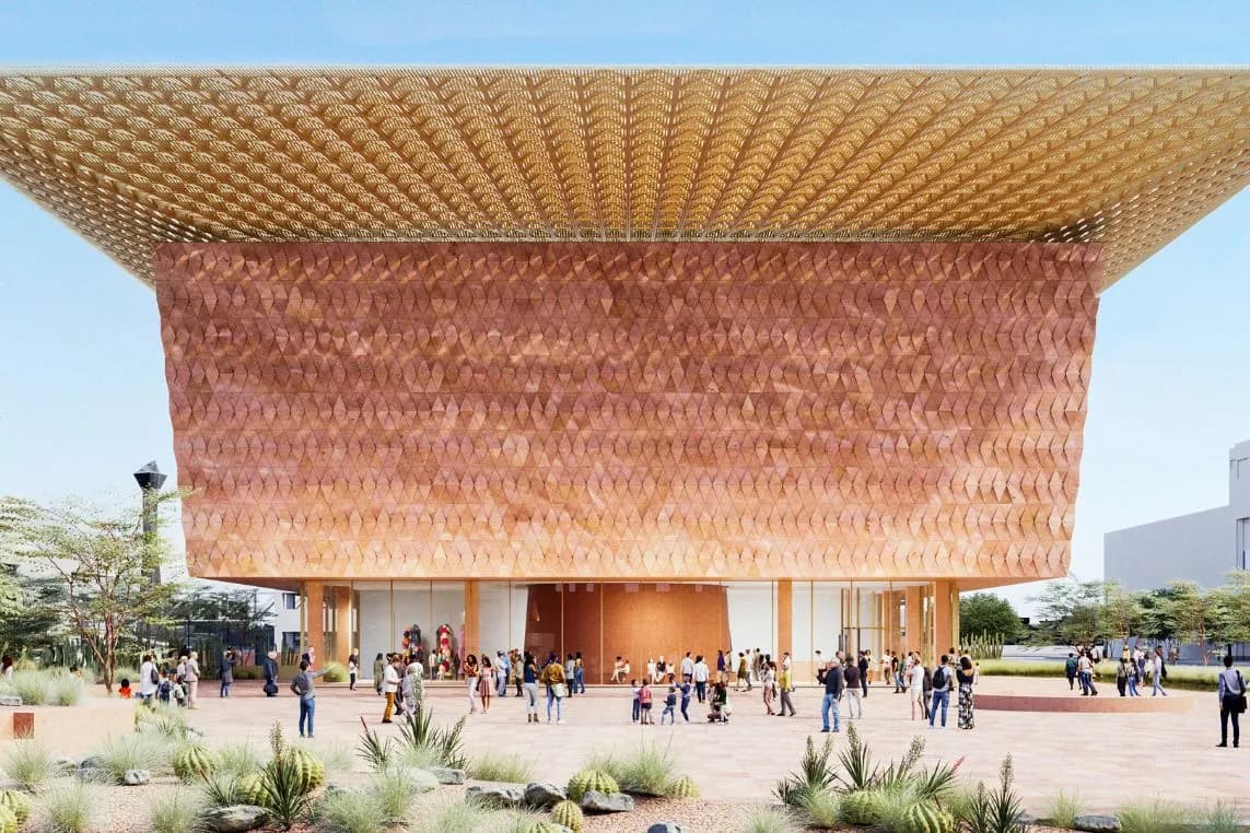 A copper-coloured building with a geometric, triangular façade and a large latticed canopy; people are gathered in a square with desert vegetation.