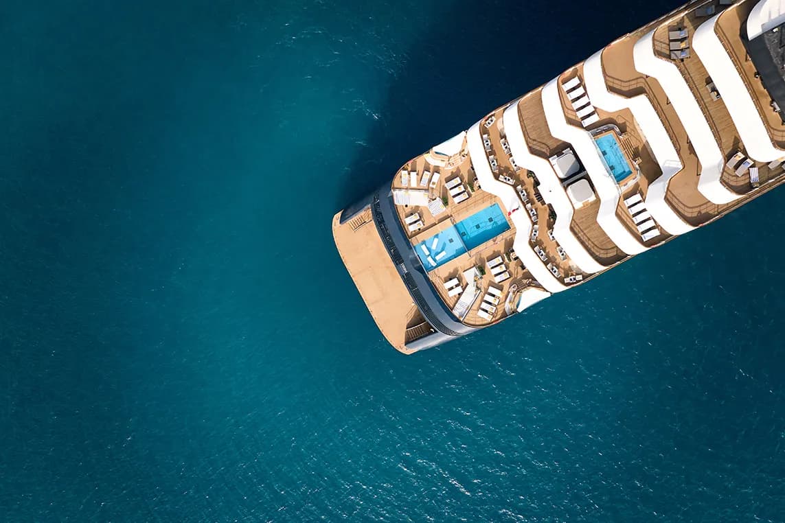 Aerial view of a luxury cruise ship deck with blue pools, sun loungers, and white curved walkways, surrounded by deep blue ocean.