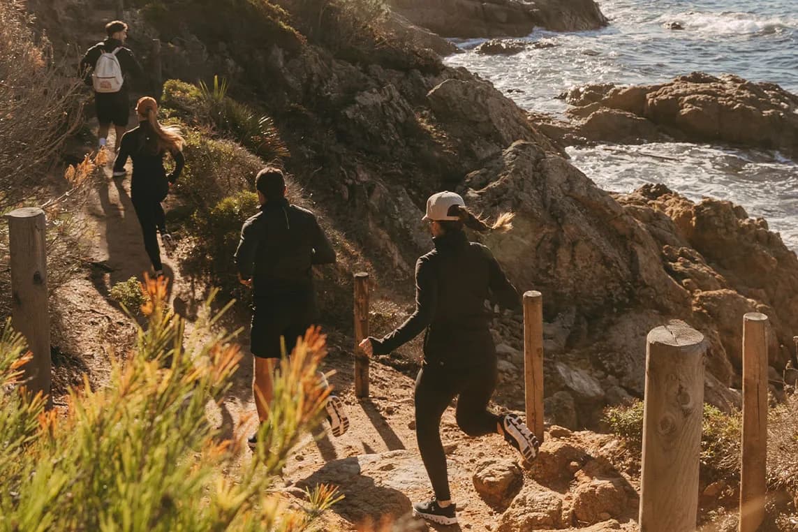 A small group of hikers walks along a rocky coastal trail with wooden posts, overlooking the sea.