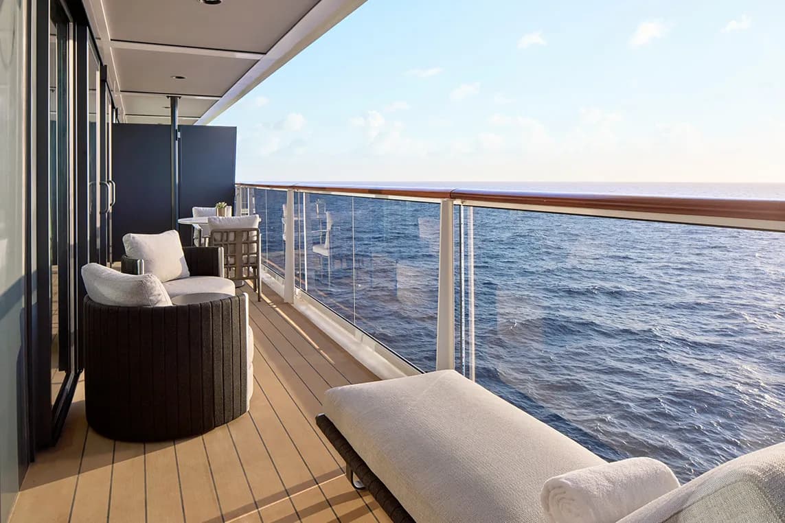 Ocean-view balcony on a cruise ship with cushioned chairs, a lounge chair, small table, and glass railing overlooking the sea.