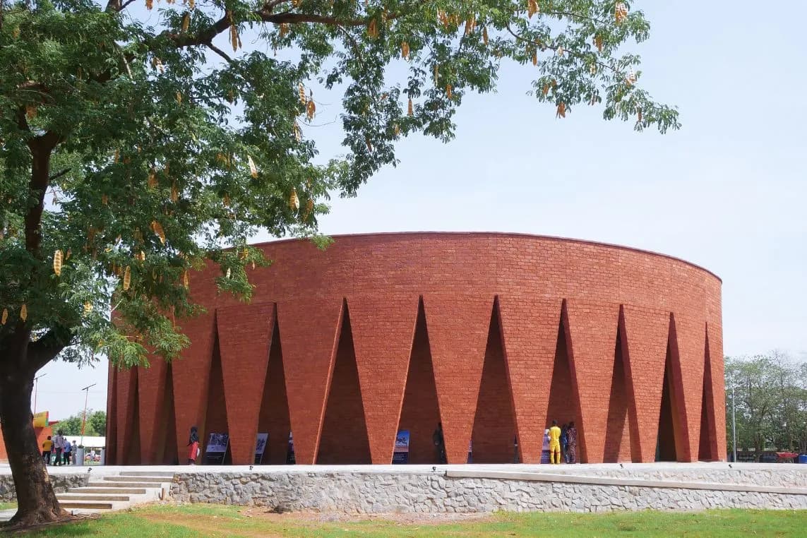 A round red-brick building with tall triangular openings at the base, standing in a park with a large, leafy tree, near which a few people are standing.