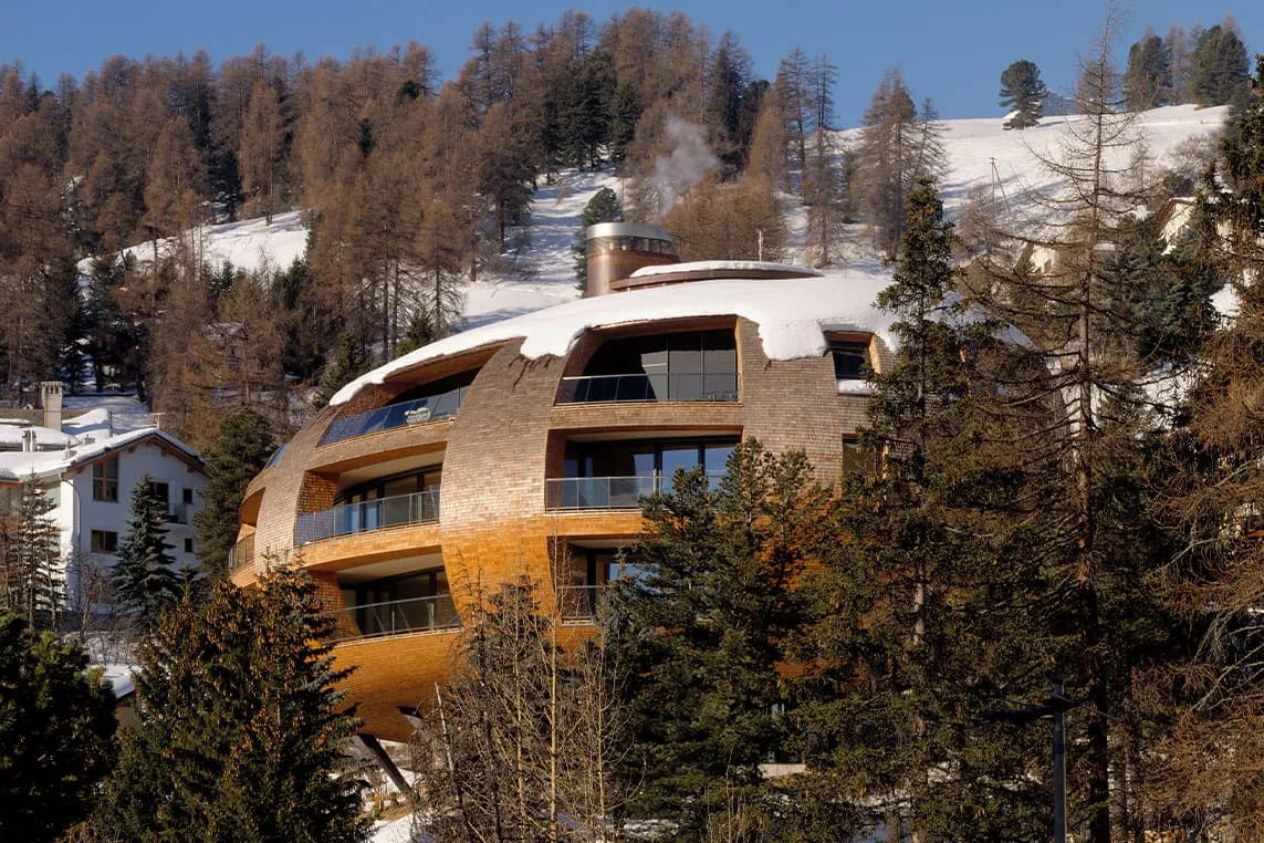 Round brick building with curved balconies sits among snow-dusted trees on a hillside; steam rises from a chimney.