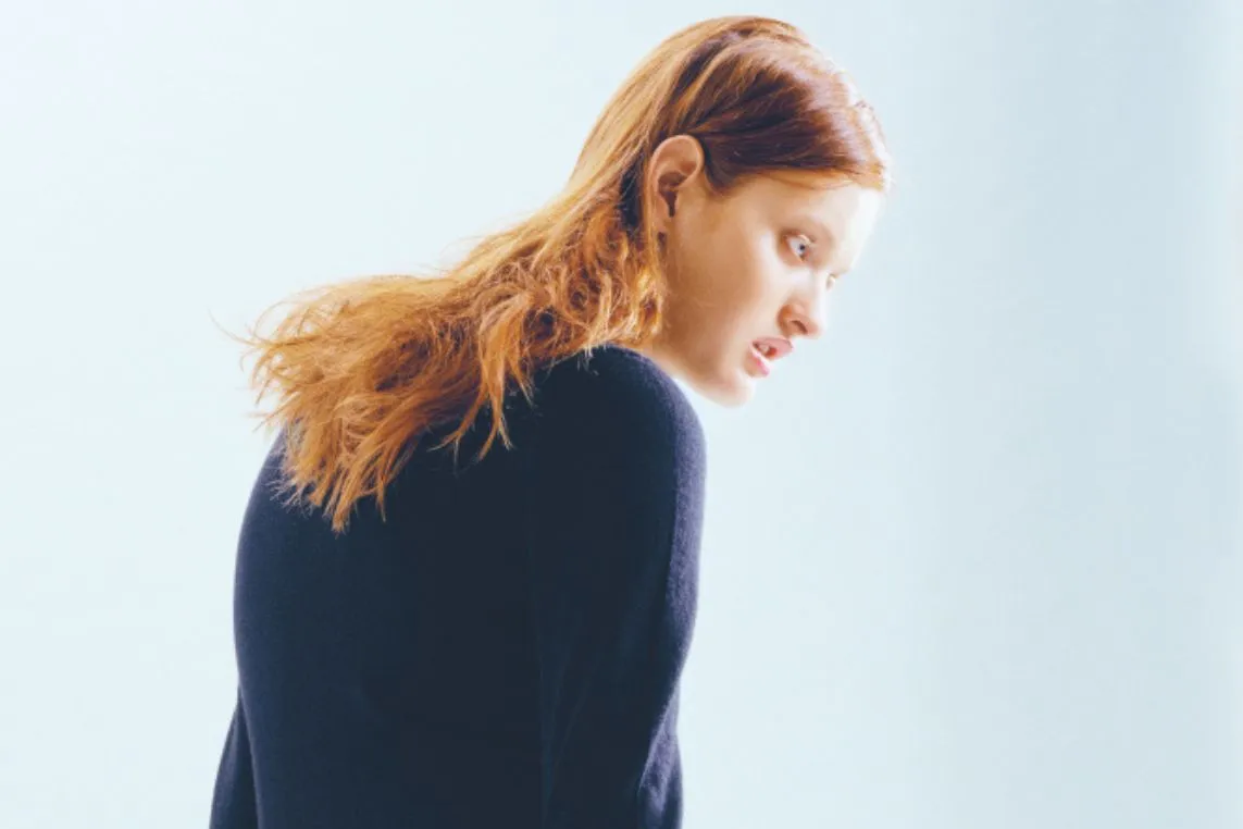 Side profile of a young woman with long red hair in a dark sweater, looking downward against a pale blue background.