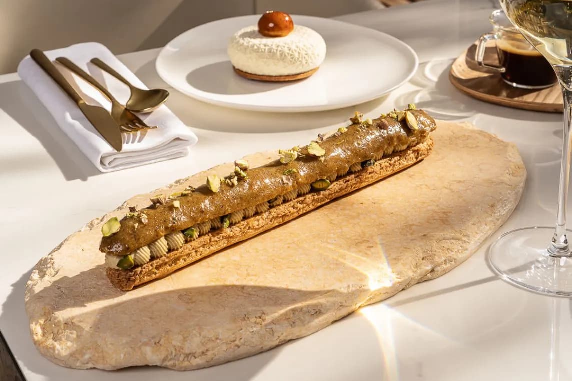 A long pastry topped with chopped pistachios rests on a flatbread on a white dining table, with a dessert, napkin, and wine nearby.