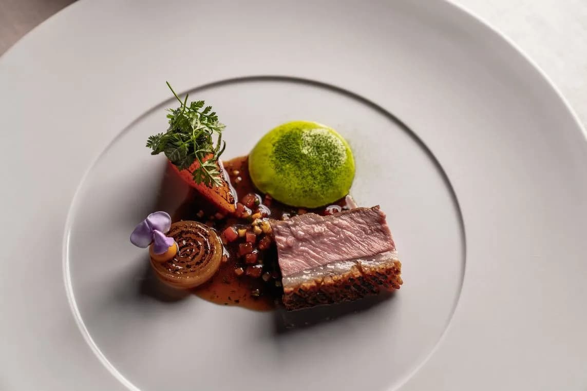Gourmet plate with seared beef slice, green sorbet ball, caramelized onion, viola flower, carrot garnish, and herbs on a white plate with sauce.
