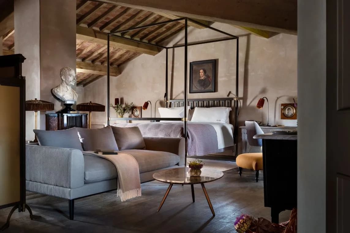 Rustic attic bedroom with a four-poster bed, gray sofa, round marble coffee table, bust sculpture, and vintage artwork.