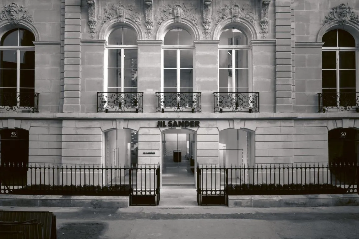 Black-and-white facade of a grand building with arched windows, ornate stonework, and a JIL SANDER entrance framed by iron gates.