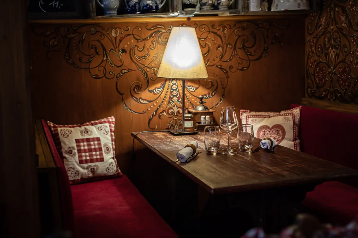 Cozy dining nook with a wooden table, red upholstered benches, heart-pattern cushions, rolled napkins, wine and water glasses, and a warm lamp.