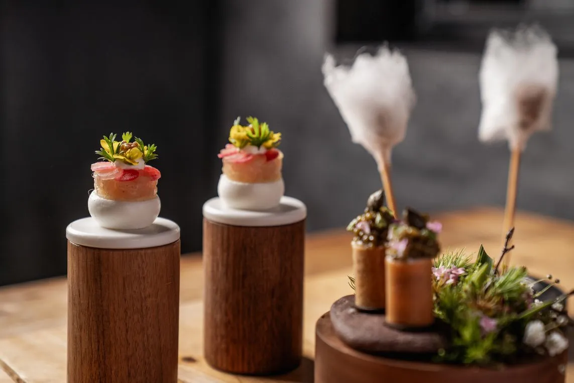Two wooden pedestals display small stacked bites topped with greens; a chocolate cake with tiny pastry cylinders and cotton candy sticks nearby.