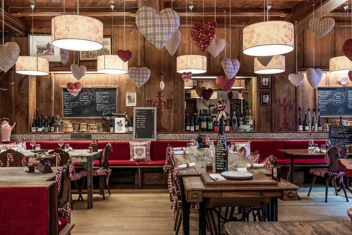 Rustic restaurant interior with wooden walls, red seating, and hanging heart-shaped decorations, warm lamps, chalkboard menus, and bottles along the bar.