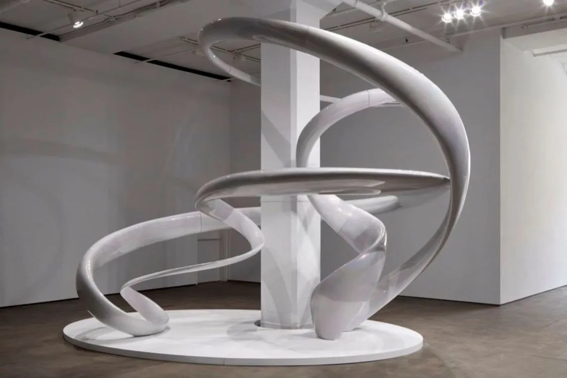 White abstract sculpture with twisting loops encircling a central pillar on a circular base in a gallery.