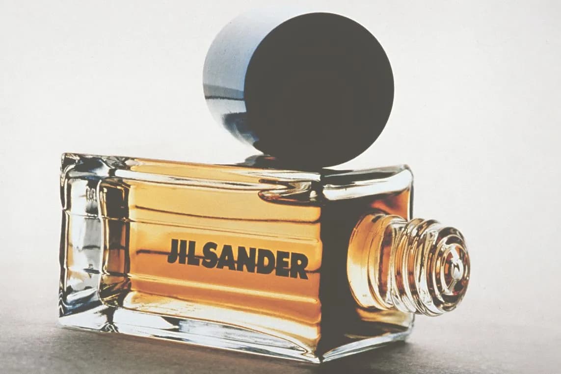 Rectangular glass perfume bottle with amber liquid and bold 'JIL SANDER' lettering, topped by a large round black cap resting on a light surface.