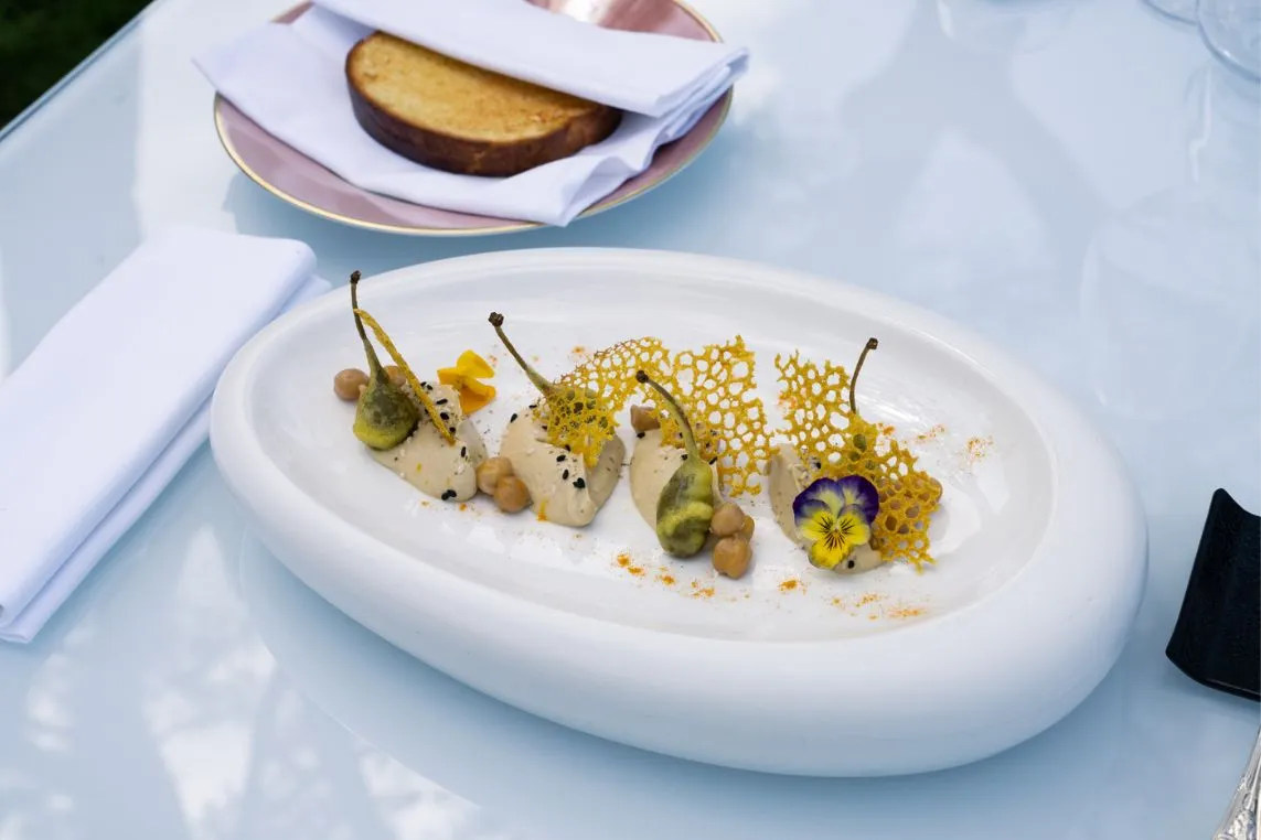 Elegant white oval plate with small bites, yellow lattice tuile, edible flowers, and nuts on a glass table; napkins and bread in background.