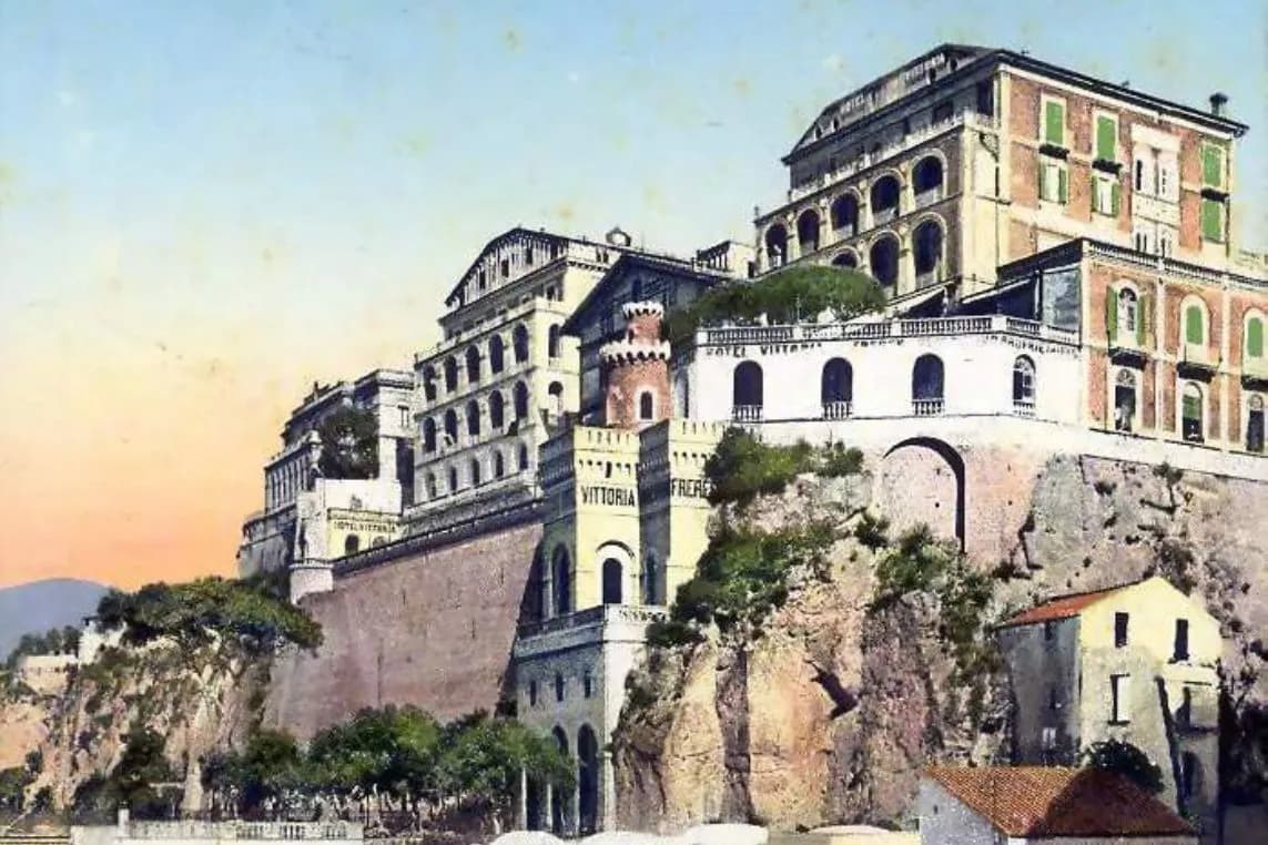 Cliffside Mediterranean villa complex with arches and terraces, a gate marked 'VITTORIA,' atop rocky cliffs at sunset.