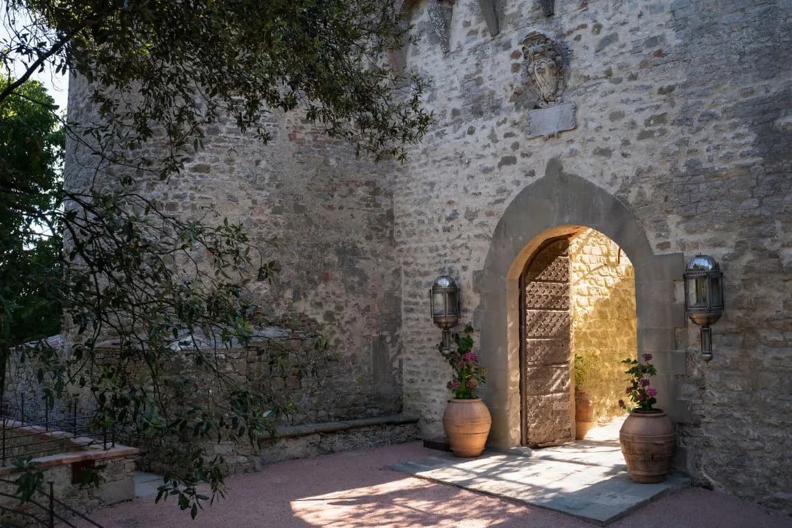 Sunlit arched stone doorway set in a historic wall, flanked by lanterns and large clay pots with flowering plants.