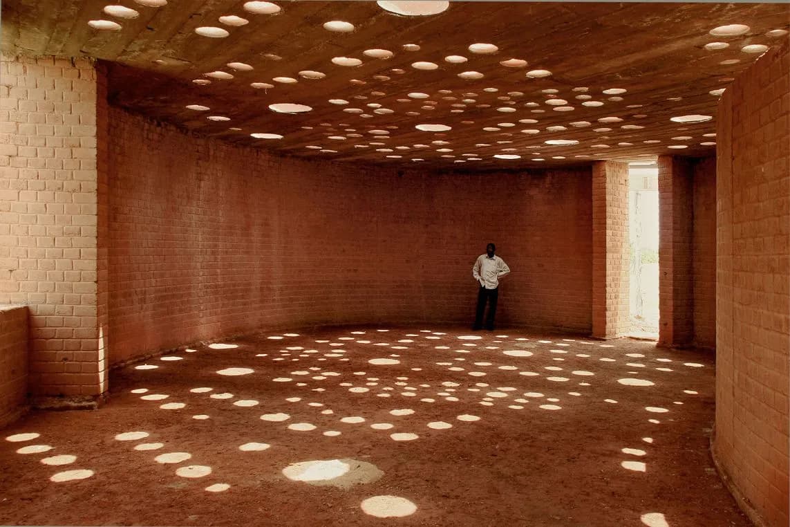 A man is standing in an unfinished brick room, into which sunlight streams through round openings in the ceiling, casting small patches of light onto the dusty floor.