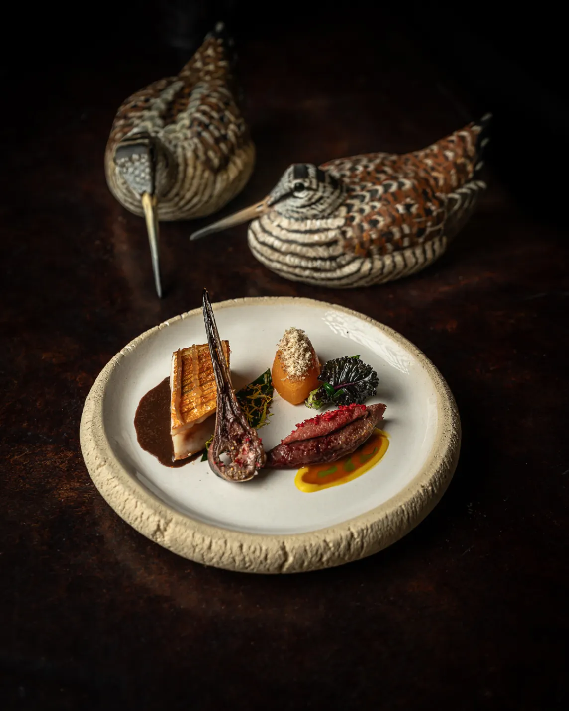 Two patterned quails loom behind an elegant plate with seared fish, meat, vegetables, and sauces on a pale ceramic dish.