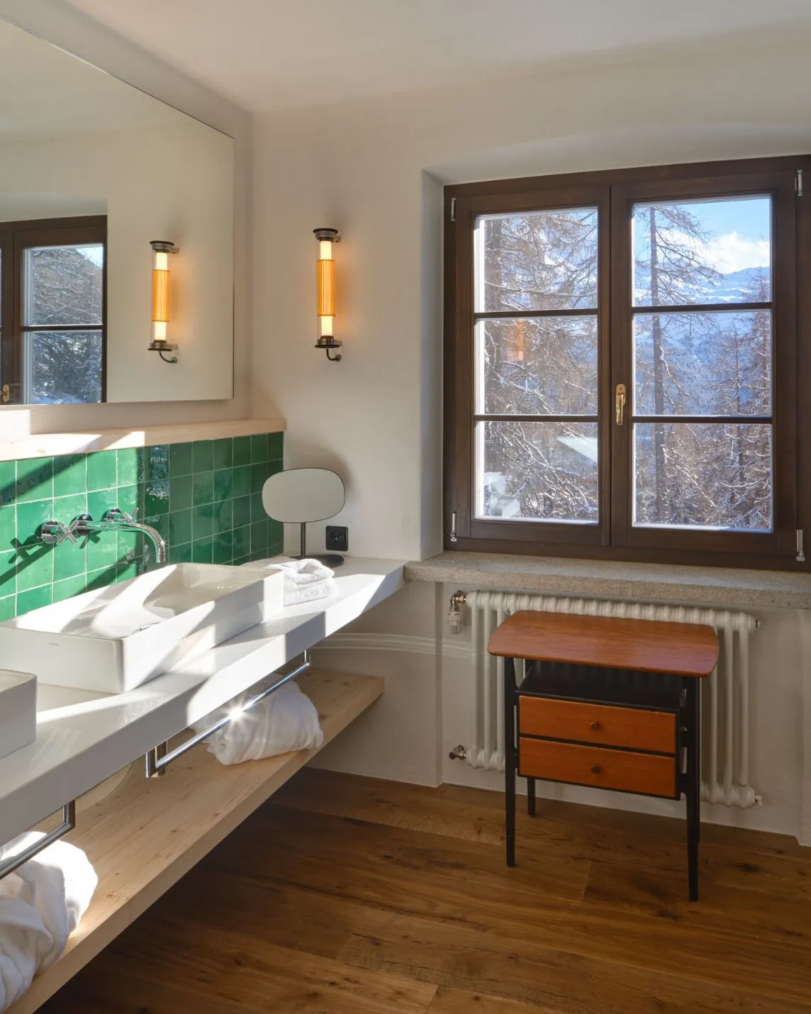 A bright bathroom with a green tiled splashback, a white washbasin, a large mirror, two wall lights and a window with a wooden frame, through which you can see snow-covered trees
