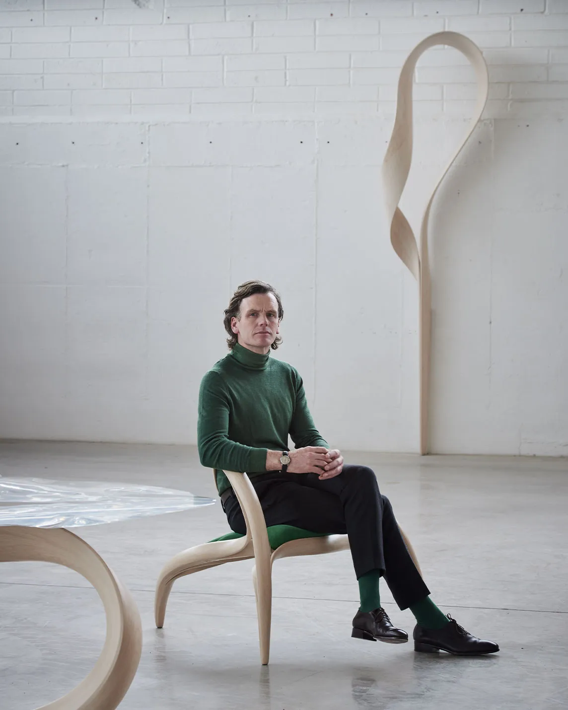 Man in a green turtleneck sits on a sculptural wooden chair in a bright white gallery, with a tall curved wood sculpture behind him.