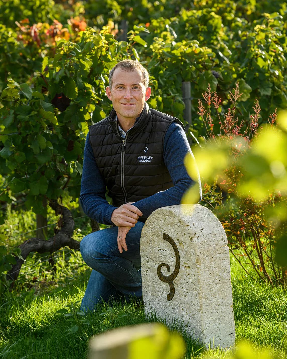 A man kneels beside a stone marker decorated with a curved pattern in a sunny vineyard. He is wearing a blue sweater and a black quilted vest.