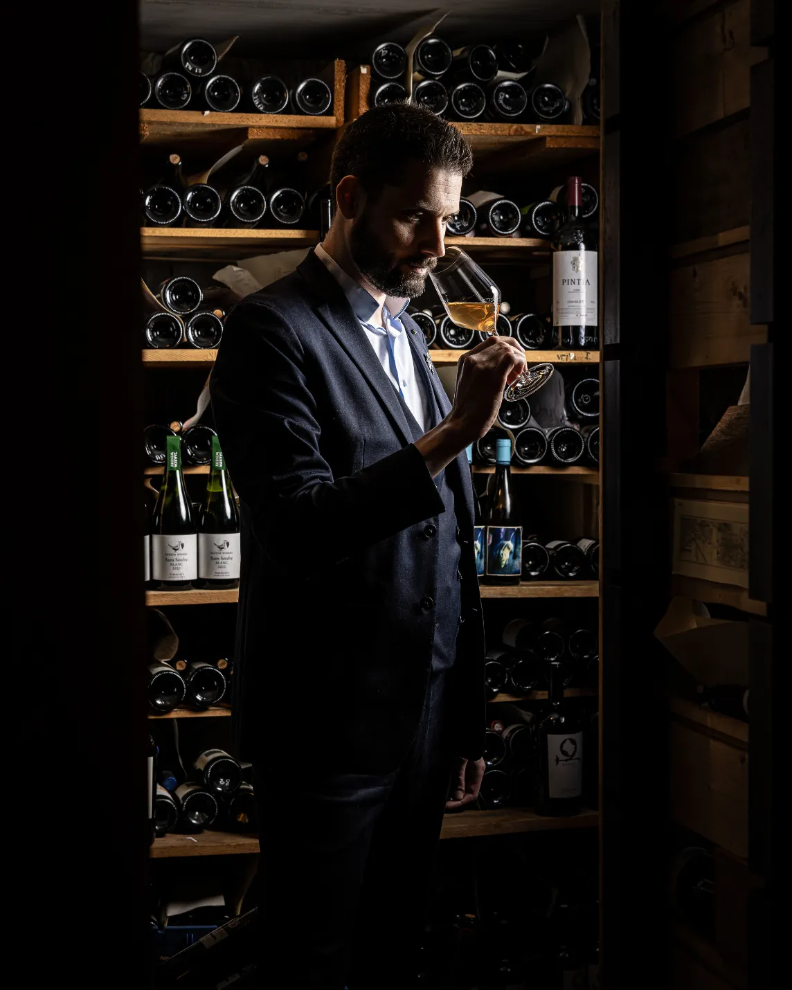 Man in a dark suit smells a wine glass in a dim wine cellar, surrounded by shelves of bottles.
