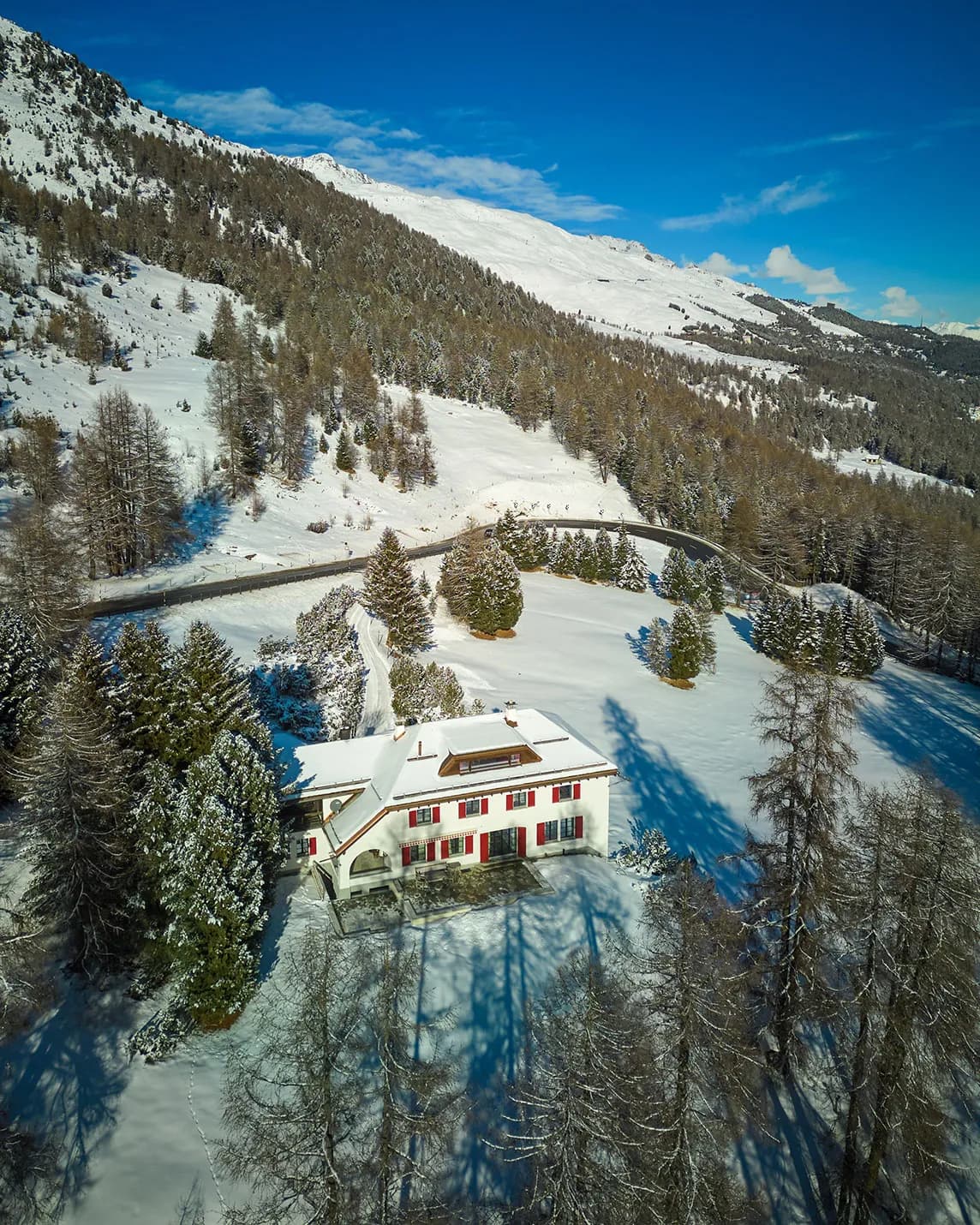An aerial view of a snow-covered mountain chalet surrounded by fir trees, a winding road and snow-covered hills beneath a bright blue sky.